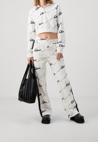Personne portant un sweat à capuche court blanc et un pantalon assorti avec l'imprimé "Colucci", tenant un sac fourre-tout plissé noir et des chaussures à plateforme noires.