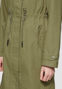 Oliwkowa kurtka z elastycznym pasem, bocznymi kieszeniami i guzikami przy mankietach. Wykonana z teksturowanej tkaniny o prostym wzornictwie.