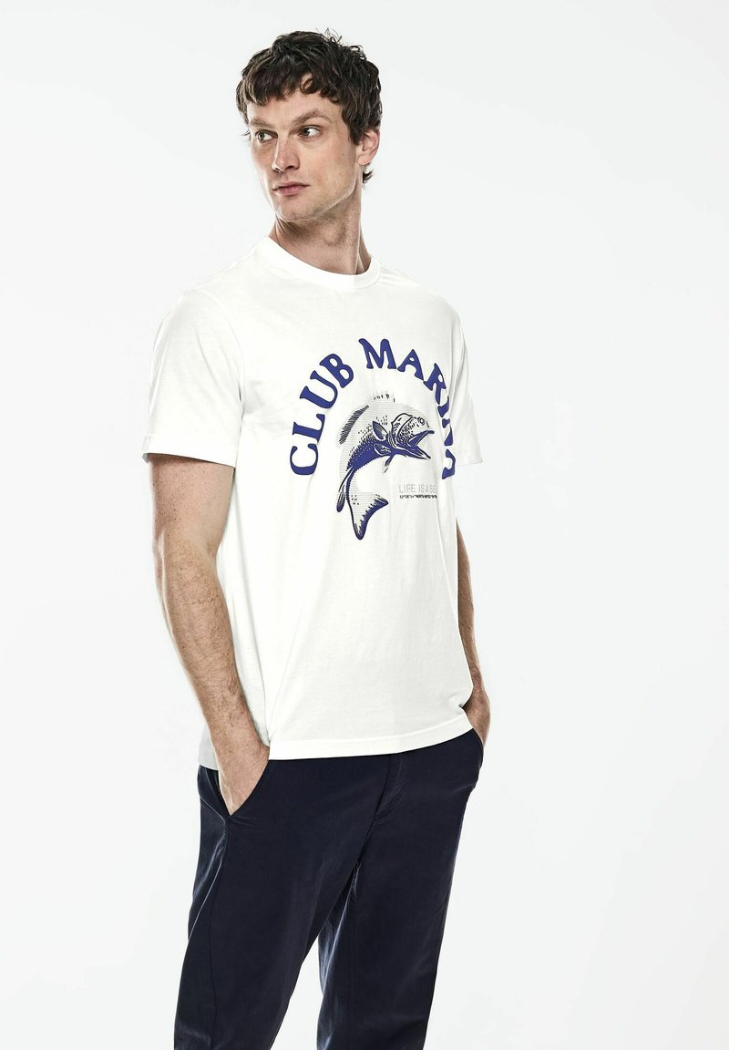 Bílá bavlněná tričko s modrou grafikou "CLUB MARINA" a ilustrací ryby, nošené s námořnickými kalhotami, s krátkými rukávy a kulatým výstřihem.