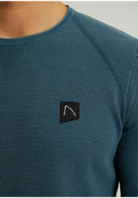 Teal textured long-sleeve shirt with a round neckline and a small black patch featuring a white abstract line design on the chest.