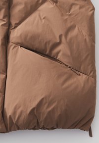 Braune Pufferjacke mit glatter Textur, diagonal gesteppt und mit einer Reißverschlusstasche an der Seite. Das Material wirkt leicht und isoliert.