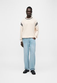 Μπεζ πουλόβερ με ναυτικές ρίγες στους ώμους, συνδυασμένο με ανοιχτό μπλε τζιν παντελόνι φαρδιάς γραμμής και μαύρα παπούτσια. Ομαλή υφή, άνετη εφαρμογή.