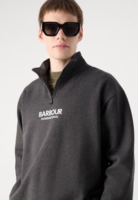 Tmavě šedá mikina s kapsami na zip a vysokým límcem, s bílým logem "BARBOUR INTERNATIONAL". Model má na sobě velké černé sluneční brýle.