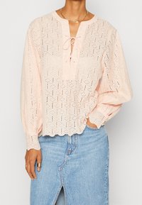Blouse en broderie anglaise rose clair avec bords festonnés et col à nouer devant, portée avec une jupe en denim bleu moyen fendue à l'avant.