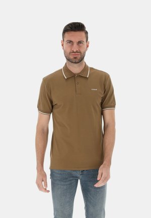 Uomo con capelli corti e scuri e barba, che indossa una polo marrone con bordi bianchi e jeans blu, in piedi davanti a uno sfondo semplice.