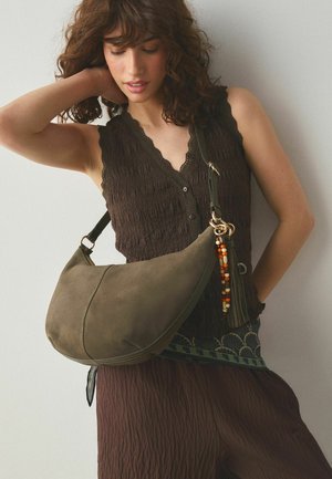 Woman with curly hair wearing a brown textured sleeveless top and pants, carrying a large olive-green shoulder bag with a tassel and beads.