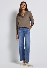 Chemise à manches longues marron clair avec un col boutonné, associée à un jean bleu à jambes larges et des baskets blanches. Tissu lisse avec une coupe décontractée.