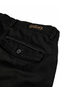 Schwarze Hose mit einer Gesäßtasche, die einen schwarzen Knopf und ein gesticktes "gallery."-Label aus goldener Fäden aufweist. Glatte Textur, maßgeschneiderter Schnitt.