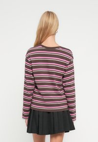 Ξανθιά γυναίκα που φαίνεται από πίσω, φοράει ένα καφέ, ροζ και λευκό ριγέ μακρυμάνικο μπλουζάκι και μια μαύρη πλισέ φούστα, σε λευκό φόντο.