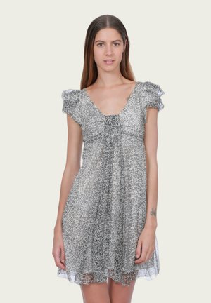 Femme aux longs cheveux bruns portant une robe courte grise à imprimé léopard avec des manches bouffantes sur un fond clair uni.