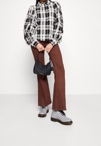 Blouse à carreaux noir et blanc avec des manches bouffantes, pantalon évasé marron, bottes grises à lacets et un petit sac à main noir. Tissus texturés et détails contrastés.
