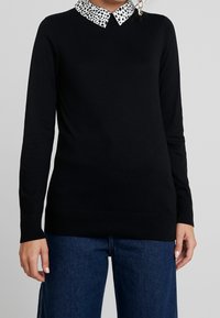 Svart långärmad tröja med en vit krage som har ett svart prickmönster. Stickat tyg med ribbad nederkant och ärmslut.