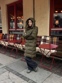 Olivengrøn dunjakke med hætte, lynlås og sidelommer. Modellen har mørke jeans og grå støvler på. Baggrunden har borde med røde toppe.