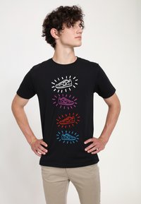 Sort t-shirt med fire sneakers grafik i hvid, lilla, rød og blå, omgivet af stråler. Kortærmet og med rund hals.