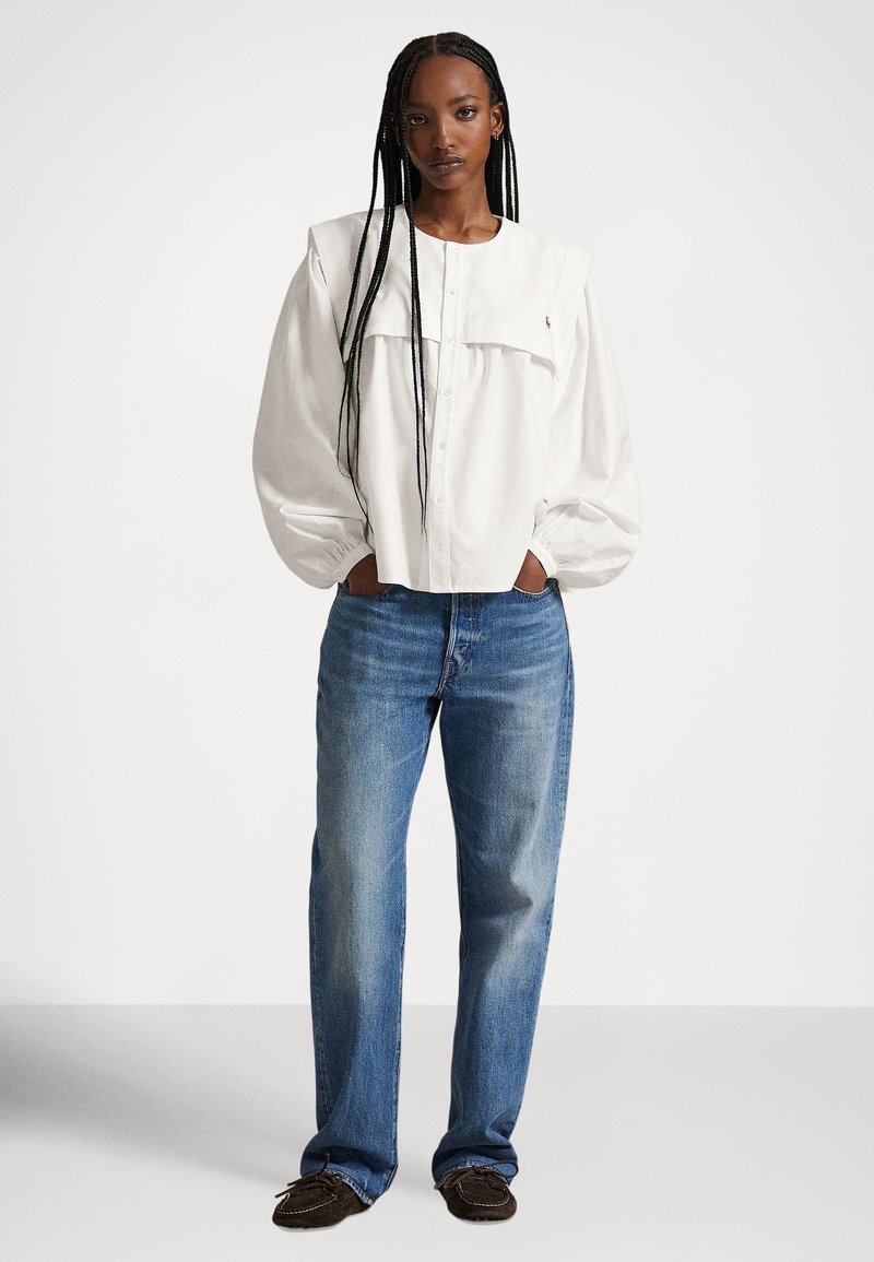 Vrouw met lange vlechten, gekleed in een wit blouse met knopen en pofmouwen, blauwe spijkerbroek en donkere mocassinschoenen, staand met handen in de zakken.