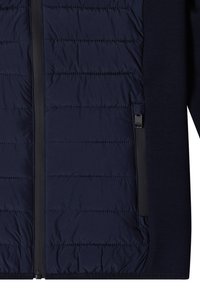 Veste matelassée bleu marine à l'avant avec des coutures horizontales, une fermeture éclair centrale et une poche latérale zippée sur le côté droit.