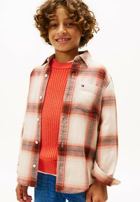 Camicia di cotone a quadri nei toni crema, arancione e marrone, sovrapposta a un maglione arancione testurizzato. Presenta chiusura con bottoni e una tasca sul petto.