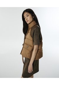 Bruine puffer vest met een hoge kraag, een voorzak met drukknoopsluiting en stoffen accenten op de schouders. Draagt over een gestructureerde bruine jurk.