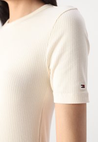Geribbelde beige T-shirt met korte mouwen, met een ronde halslijn en een klein logo in rood, wit en blauw op de manchetten. Zachte textuur.