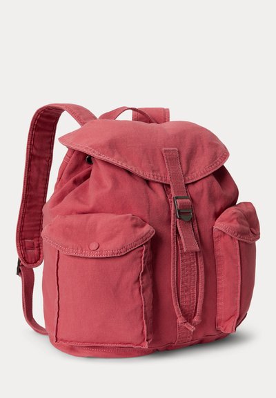 Κόκκινο υφασμάτινο σακίδιο με δύο μπροστινές τσέπες, κλείσιμο με καπάκι που ασφαλίζει με λουράκι και μεταλλικό κούμπωμα, και ρυθμιζόμενους ώμους.