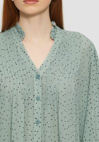 Jasnozielona bluzka z falbaną przy kołnierzyku, wykonana z teksturowanej tkaniny w czarne kropki. Zawiera dekolt w kształcie litery V oraz zapięcie na guziki.