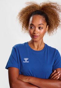 Blauwe katoenen t-shirt met een ronde hals en korte mouwen. Witte geborduurde logo op de linkerkant van de borst. Glad oppervlak overal.