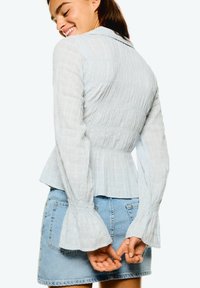 Lichtblauw, textuur blouse met een peplum taille, lange klokmouwen en een kraag, gecombineerd met een jeansrok. Details van subtiel patroon zichtbaar.
