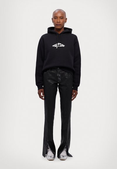 Schwarzer Hoodie mit weißem Logo, kombiniert mit glänzenden schwarzen weiten Hosen mit Seitenschlitzen und silbernen Schuhen mit spitzen Zehen.