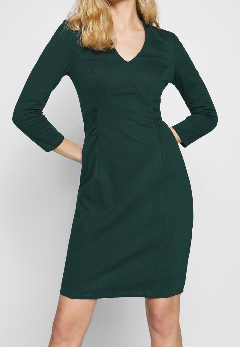Femme portant une robe ajustée vert foncé, longueur genou, avec manches trois-quarts et un décolleté en V, posant les mains sur les hanches.