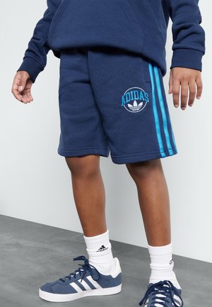 Osoba nosząca granatowe szorty Adidas z jasnoniebieskimi paskami, pasującą bluzę z kapturem, białe skarpetki Adidas oraz granatowe buty sportowe Adidas.