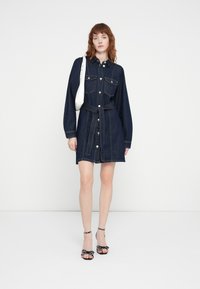 Robe chemise en denim bleu foncé, avec des manches longues, deux poches poitrine, une taille ceinturée et une fermeture à boutons sur le devant. Associée à des sandales noires et blanches.