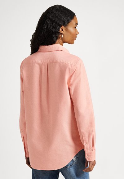 Gaiši rozā garroku krekls ar apkakli, pogu aizdari un aizmugurējo līplokšķēlumu. Mīksta tekstūra, noapaļots apakšmala un aproču aizdare ar pogām. Kombinēts ar džinsa materiālu.