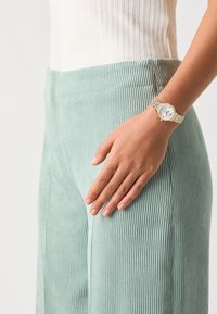 Mint green corduroy trousers with vertical ribbed texture, paired with a silver mesh watch featuring a white face and small hour markers.