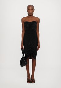 Svart strapless klänning med rynkningar, längd till mitten av låret och sidoklyvning. Modellen håller en svart kuvertväska; skorna har tunna remmar.