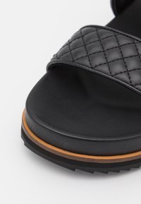 Czarne klapki z pikowaną paskiem, teksturowaną powierzchnią, wyściełaną podeszwą oraz kontrastującą brązową podeszwą.