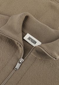 Beige Zip-Jacke mit hohem Kragen, aus strukturiertem Stoff. Verfügt über einen Metallreißverschluss und ein Etikett mit "DRYKORN" in Schwarz.