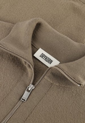 Beige Zip-Jacke mit hohem Kragen, aus strukturiertem Stoff. Verfügt über einen Metallreißverschluss und ein Etikett mit "DRYKORN" in Schwarz.
