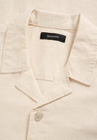 Col de chemise en coton beige avec un seul bouton et une étiquette noire portant l'inscription "Matinique" cousue à l'intérieur de l'encolure.
