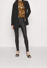 Blazer noir ajusté, blouse marron à motifs floraux, pantalon skinny noir en simili cuir, et sandales à talons noires avec détails tressés.