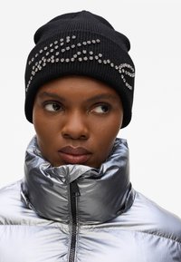 Gorro de punto negro con adornos de strass en la parte frontal, combinado con una chaqueta acolchada plateada brillante con cuello alto.