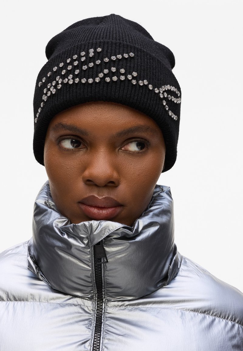 Gorro de punto negro con adornos de strass en la parte frontal, combinado con una chaqueta acolchada plateada brillante con cuello alto.