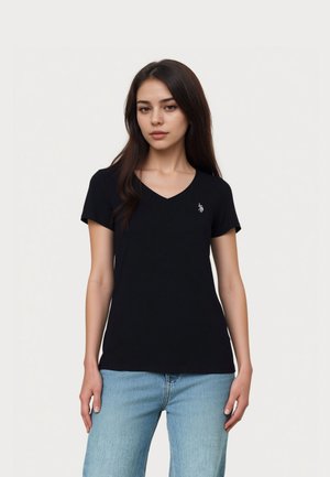 Mladá žena s dlhými tmavými vlasmi, oblečená v čiernom tričku s výstrihom do V a svetlomodrých džínsoch, stojaca pred obyčajným svetlým pozadím.