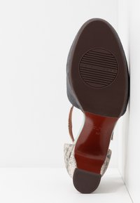 Semelle d'une chaussure à talon haut avec talon et avant en caoutchouc texturé brun foncé, dessus en similicuir à motif peau de serpent beige, photographiée sur fond blanc.