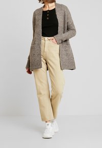 Grijze gebreide cardigan met lange mouwen, open voorkant en zijzakken over een zwarte ribbeltop. Beige recht-pijpenbroek en witte sneakers.