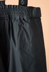 Pantalon noir taille haute avec ceinture élastique et sangle noire réglable sur un fond pêche pâle.