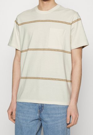 Mand iført en beige T-shirt med korte ærmer med orange vandrette syninger og en brystlomme, kombineret med blå jeans.