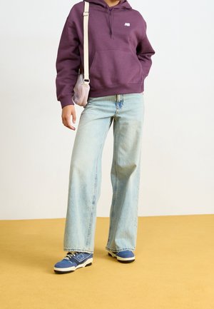 Personne portant un sweat à capuche violet, un jean large bleu clair, des baskets bleues et portant un sac à bandoulière rose clair sur un sol jaune.