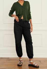 Cardigan verde a maglia con scollatura a V e chiusura a bottoni, abbinato a pantaloni cargo neri con polsini elasticizzati e sandali con cinturini argentati.