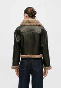 Giacca corta in pelle marrone con collo e orlo in pelliccia color cognac, caratterizzata da fibbie laterali e una texture liscia. Indossata con jeans scuri in denim.