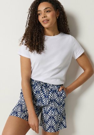 Kobieta z kręconymi włosami ubrana w białą koszulę z krótkim rękawem i granatowe spodenki w biały wzór liści, stojąca z ręką w kieszeni.